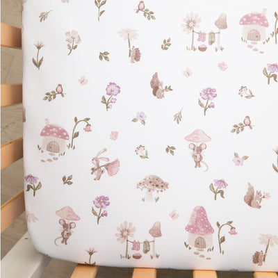 Close-up overhead view of the Cotton Jersey Fitted Sheet in Mushroom House inside a wooden crib, showcasing a whimsical pattern of pink mushroom cottages, little forest mice, and scattered wildflowers on a crisp white background.