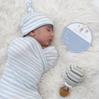 Close-up birth announcement shot of a sleeping newborn baby swaddled tightly in a light blue and white striped jersey blanket and matching top-knot beanie, resting on a white faux fur rug next to a write-in milestone disc and a knit hot air balloon rattle.