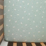 Close-up overhead view of the Cotton Jersey Fitted Sheet in Little Geese, showing a cute pattern of mini white ducks and tiny yellow flowers on a soft sage green baby mattress cover.