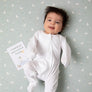 Smiling baby in a white onesie lying flat on a sage green Little Geese printed crib sheet holding a milestone card.
