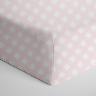 Detailed close-up studio view of the deep-pocket corner seam of a 100% cotton jersey fitted crib sheet featuring a classic pale pink and white gingham checkered pattern.