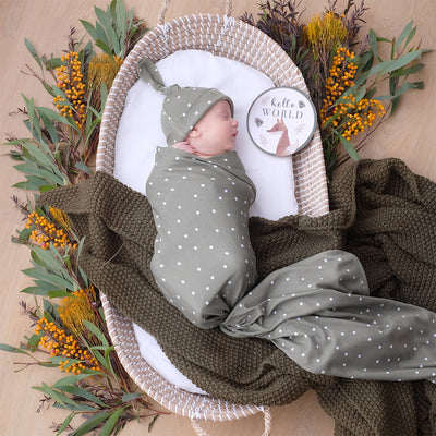 Overhead birth announcement portrait of a sleeping newborn swaddled tightly in an olive green jersey blanket and matching knotted hat with a white polka dot pattern, resting in a woven basket styled with autumn eucalyptus florals and an olive knit blanket.
