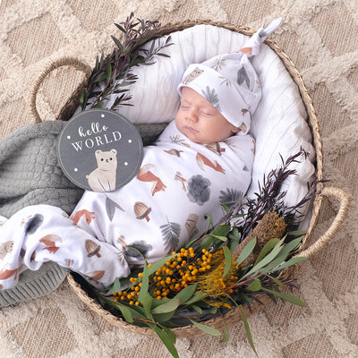 Overhead birth announcement portrait of a sleeping newborn swaddled tightly in a white jersey wrap and matching knotted hat with a woodland animal print, resting in a woven basket styled with deep green eucalyptus, rustic yellow flowers, and a dark charcoal bear milestone card.