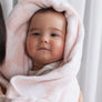 A cute baby looking at the camera with a soft Pink Check Knitted Chenille Blanket draped over their head like a hood.