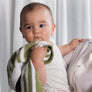 Cute baby holding and wrapped in a soft Knitted Chenille Blanket in Sage Stripes against a soft-focus background.