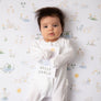 Baby wearing a white onesie lying flat on a soft Goosey Goosey Cotton Jersey Fitted Sheet while holding a nursery milestone card.
