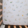 Close-up overhead view of the Cotton Jersey Fitted Sheet in Goosey Goosey inside a crib, featuring an illustrated pattern of white geese, little ponds, frogs, and yellow suns.