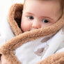 Close-up of a baby bundled up warmly, showing the thick, soft brown sherpa trim of the Cuddly Bear baby blanket framed around the baby's face.