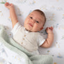 Happy baby lying down and covered comfortably with the cozy Goosey Goosey Jersey Sherpa Blanket on a matching printed crib sheet.